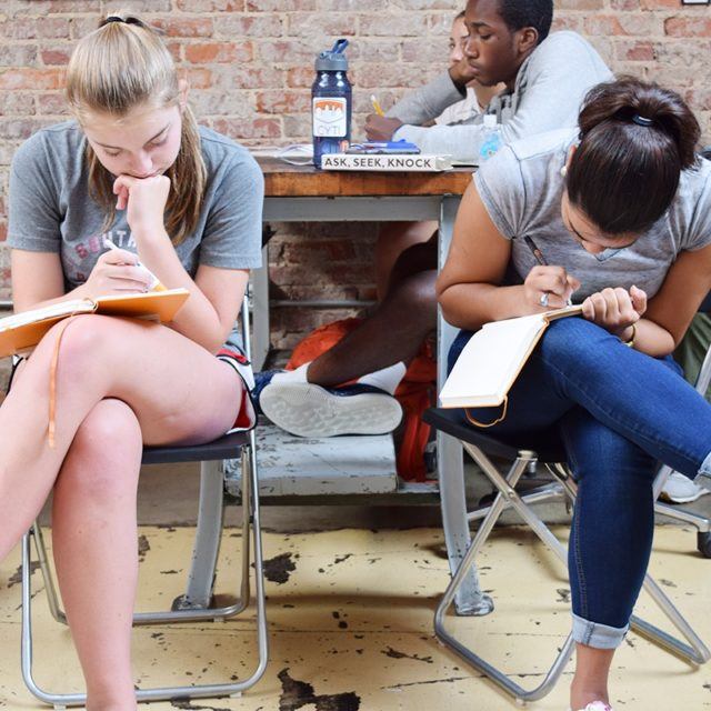 Two girls are seated while writing in notebooks