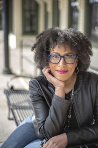 Headshot of black female with curly chin length hair wearing glasses and red lipstick. She is leaning her chin on her hand.