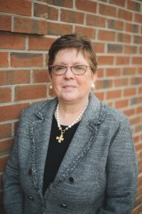 White female with short hair wearing glasses and a grey blazer. She is standing against a brick background.
