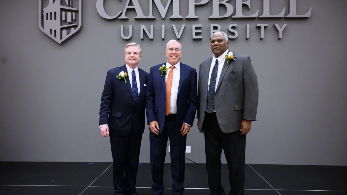 Three individuals in suits standing and pose for a photo. Campbell University and its logo of Kivett Hall are in the background on the wall behind the three individuals.
