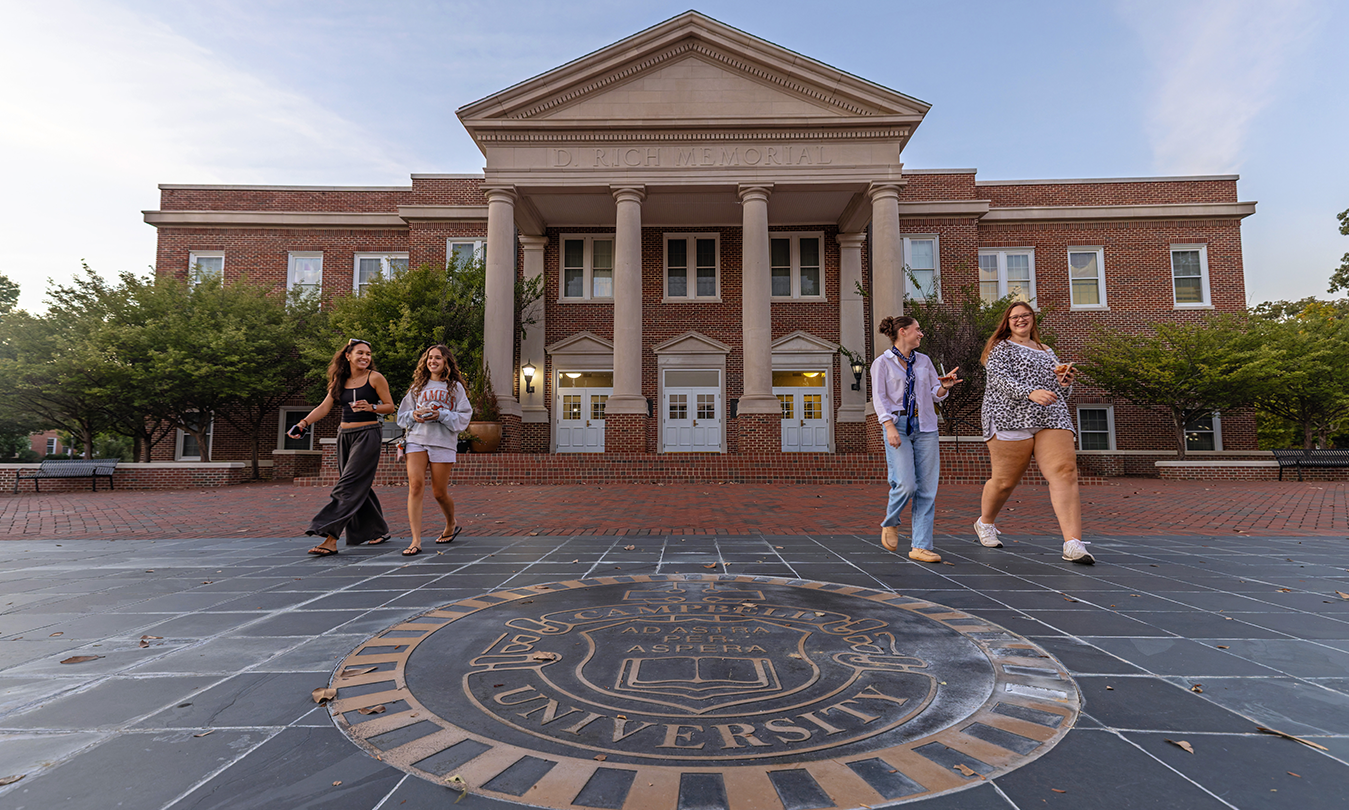A group of people walk in front of a large brick building with columns at Campbell University. The university emblem is displayed on the stone pavement in the foreground.