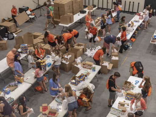 an overhead shot of a large room full of tables, at which students wearing orange are packing boxes full of supplies