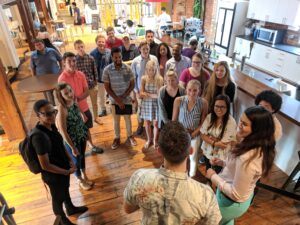 An overhead view of 25 students gathered around Tom Jacobs in the HQ workspace. 