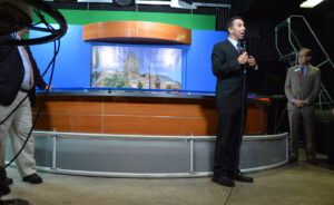 Professor Brian Bowman stands in front of the new wooden and metallic newsroom desk, speaking to an audience in a black suit. 