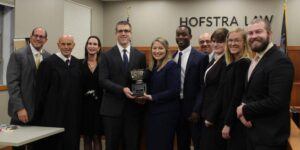 Photo of five Campbell Law advocates holding national trophy in courtroom under Hofstra Law sign with judges