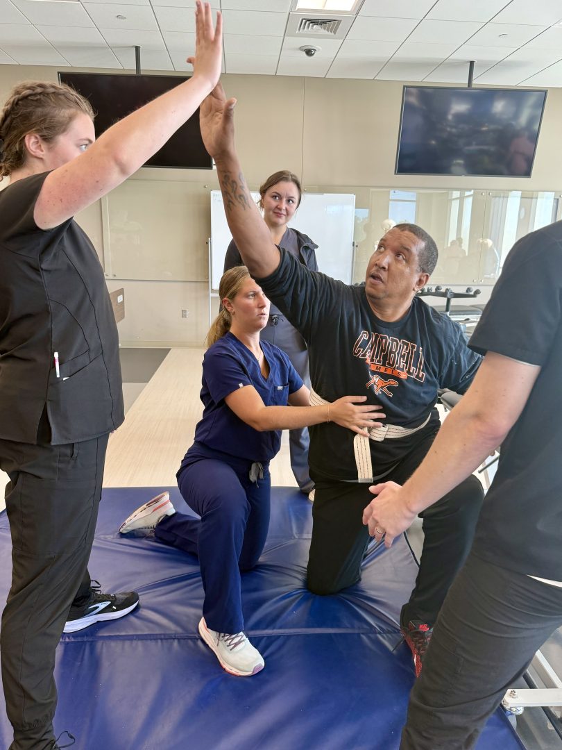 Patient reaches up to touch student's hand while Anna Fish stabilizes the patient.