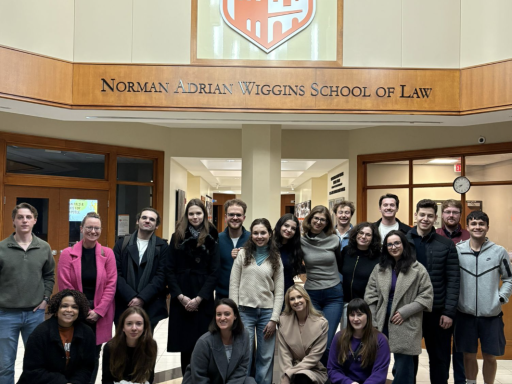 Photo of 12 Romanian students posing along with Campbell Law students in the lobby of the law school under the sign that reads Norman Adrian Wiggins School of Law and the orange and white Civet