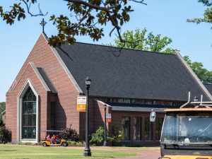 Butler Chapel at Campbell University, featuring red brick architecture, a large Gothic-style front window and a cross atop its roof. A golf cart drives past the chapel on a sunny day beneath tree branches.