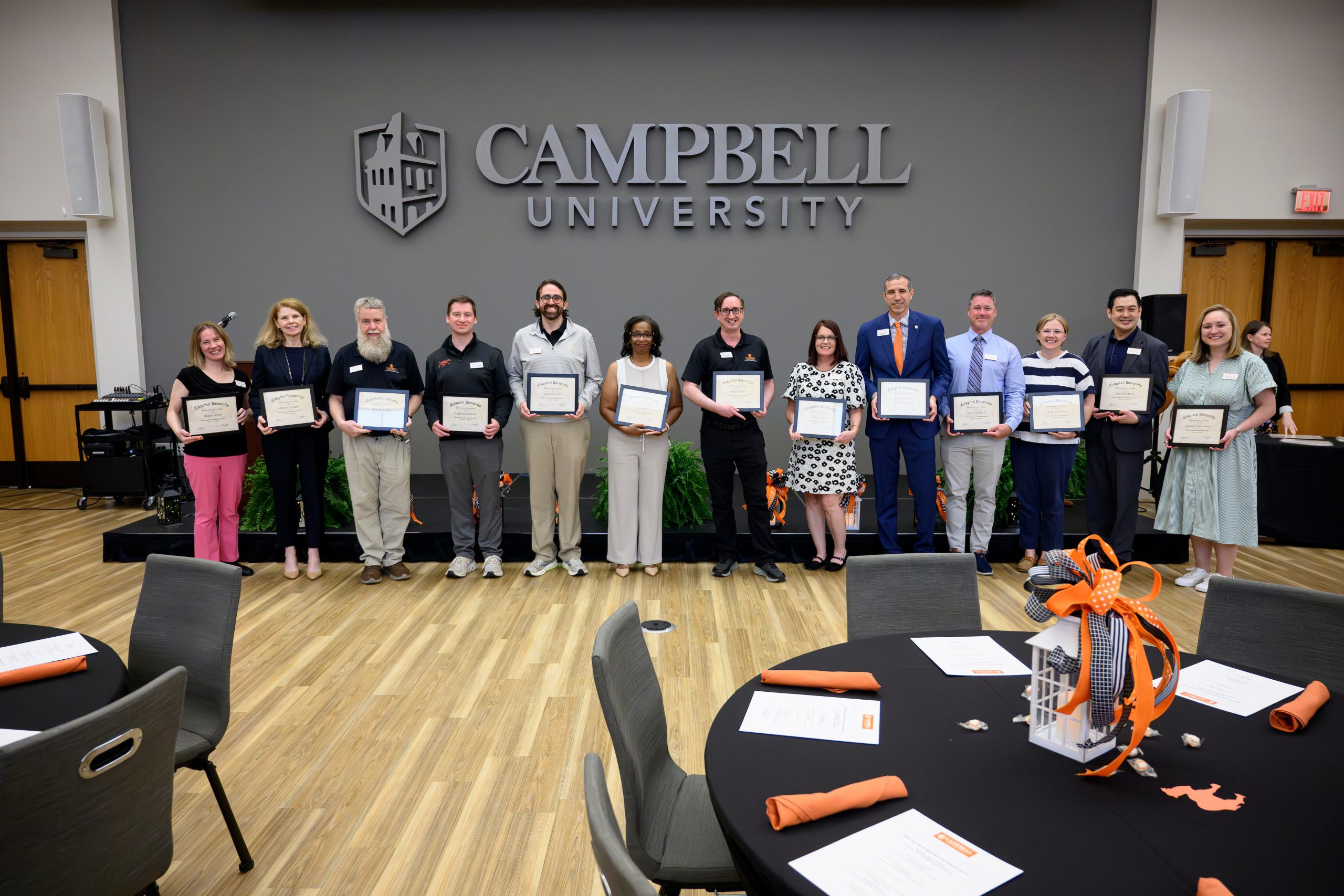 Male and female employees with 5 years of service each holding their certificate standing in front of a low stage in the student union ballroom.