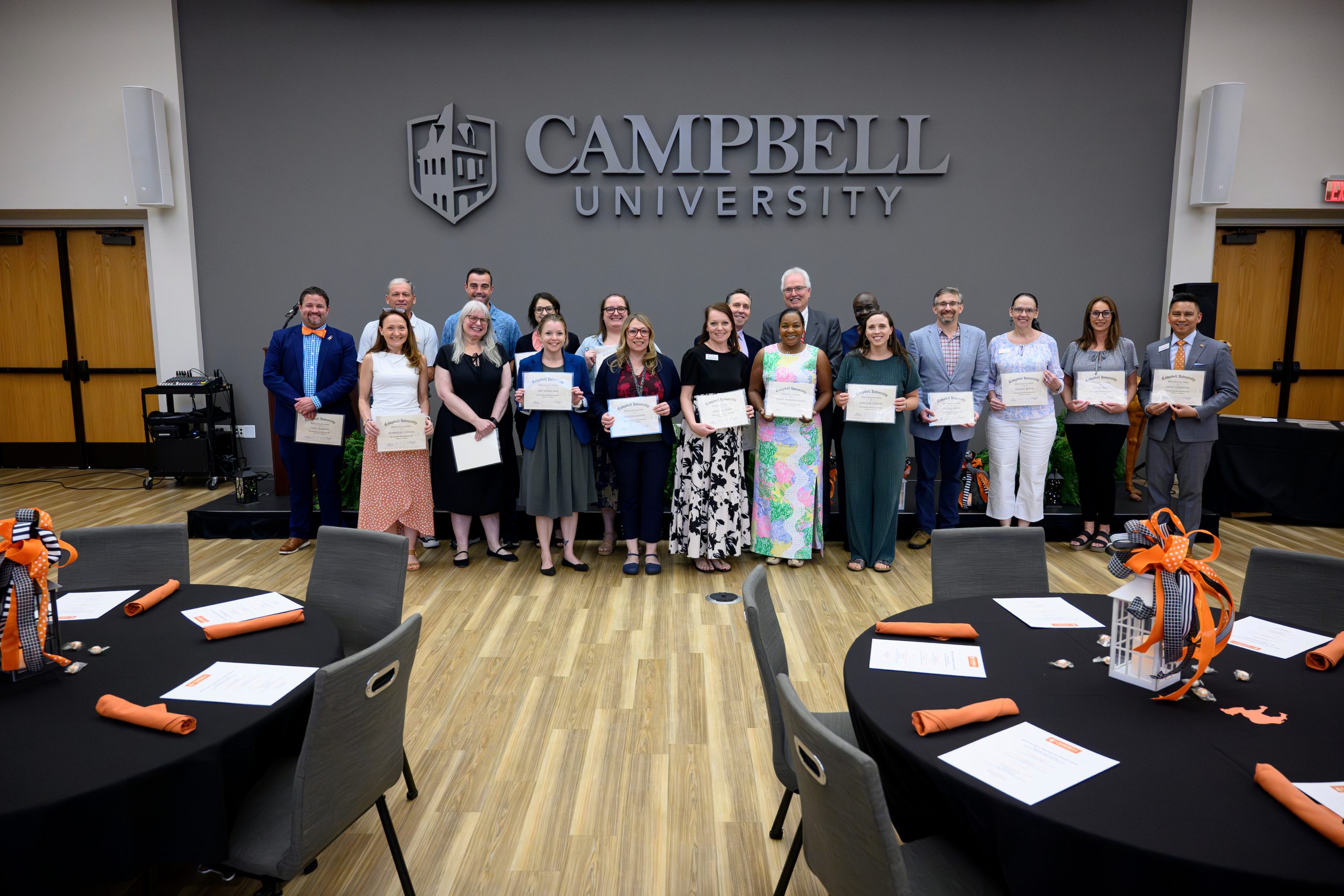 Male and female employees with 10 years of service each holding their certificate standing in front of a low stage in the student union ballroom.