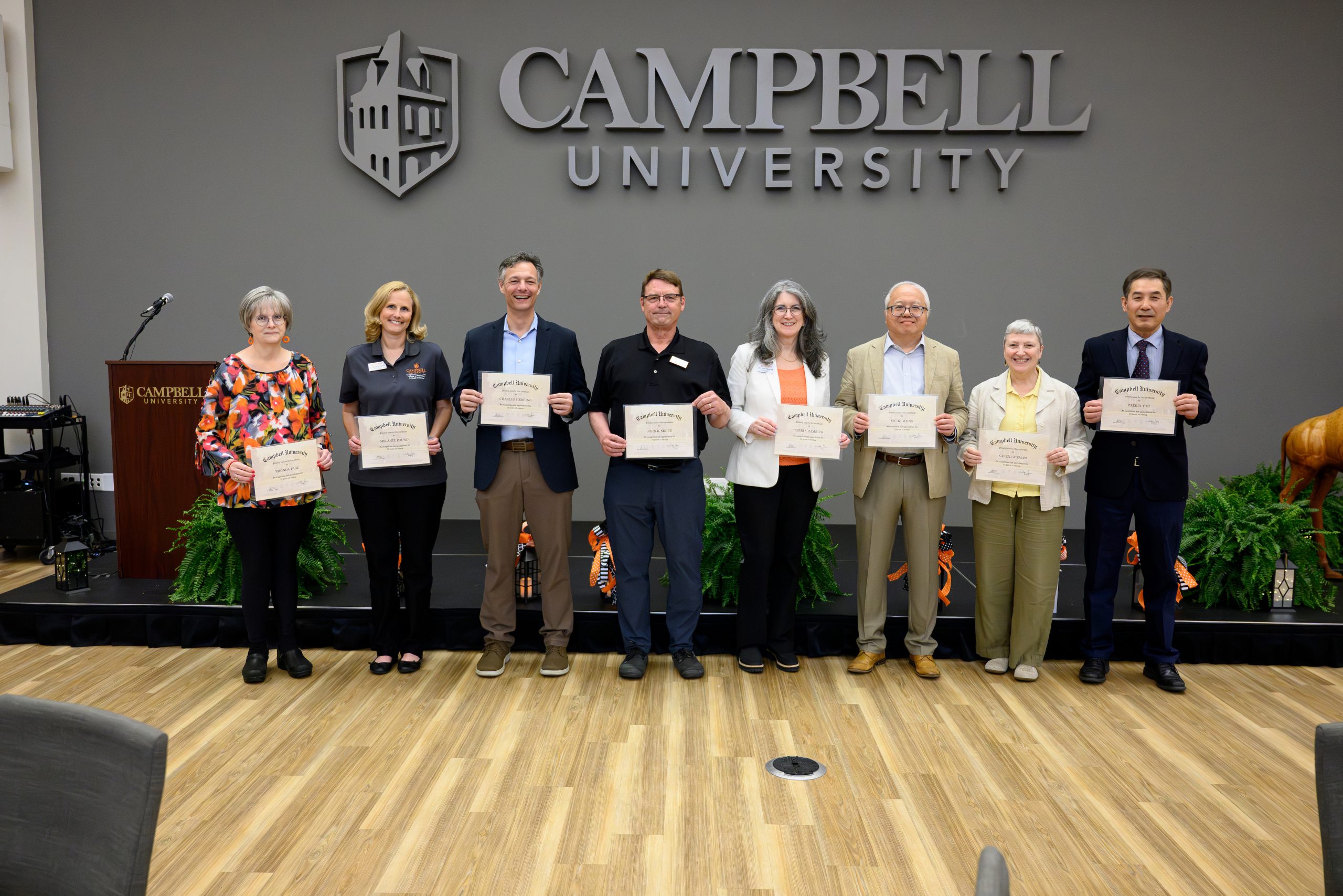 Male and female employees with 25 years of service each holding their certificate standing in front of a low stage in the student union ballroom.