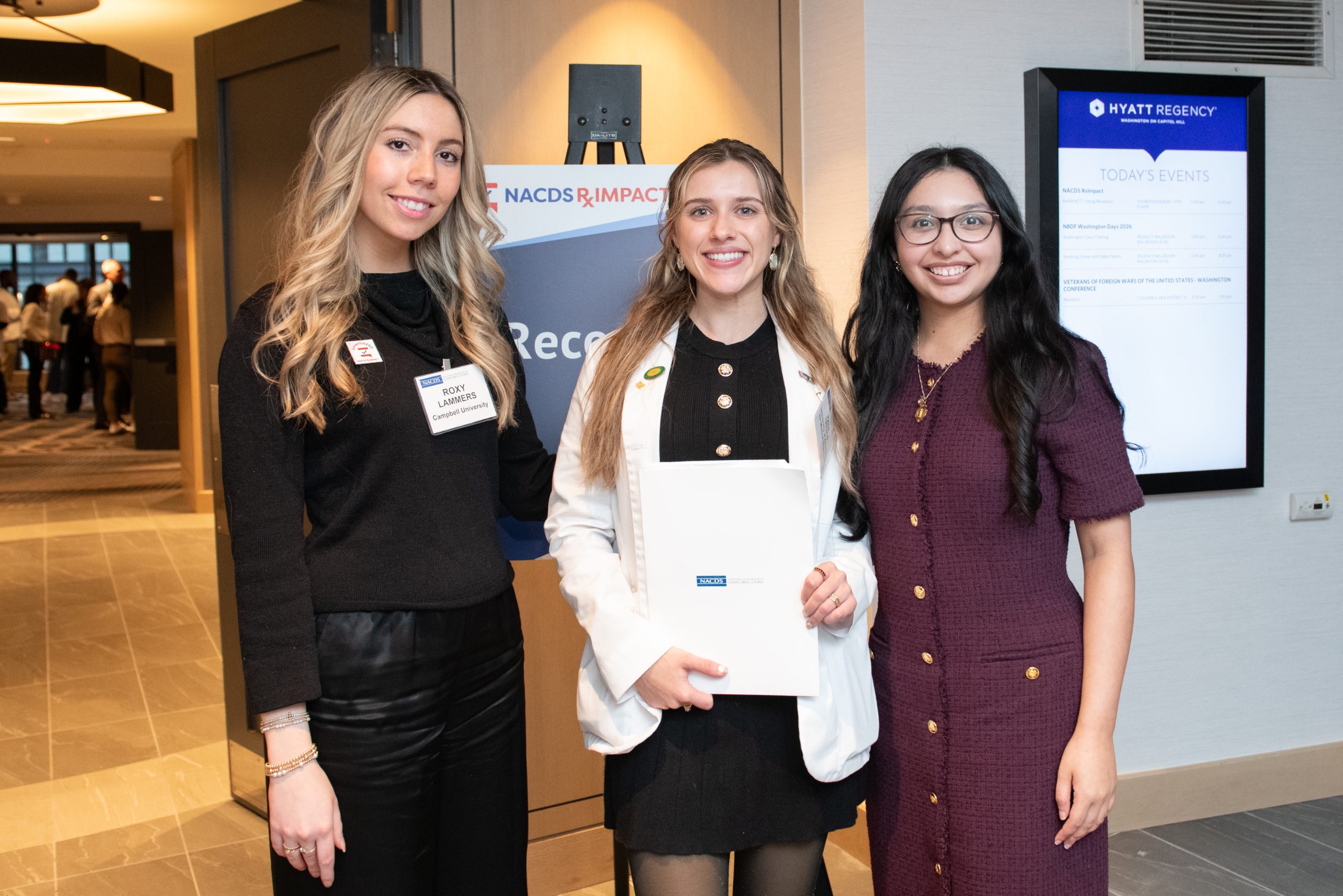 Three female students wearing professional attire standing together outside a conference room of a hotel