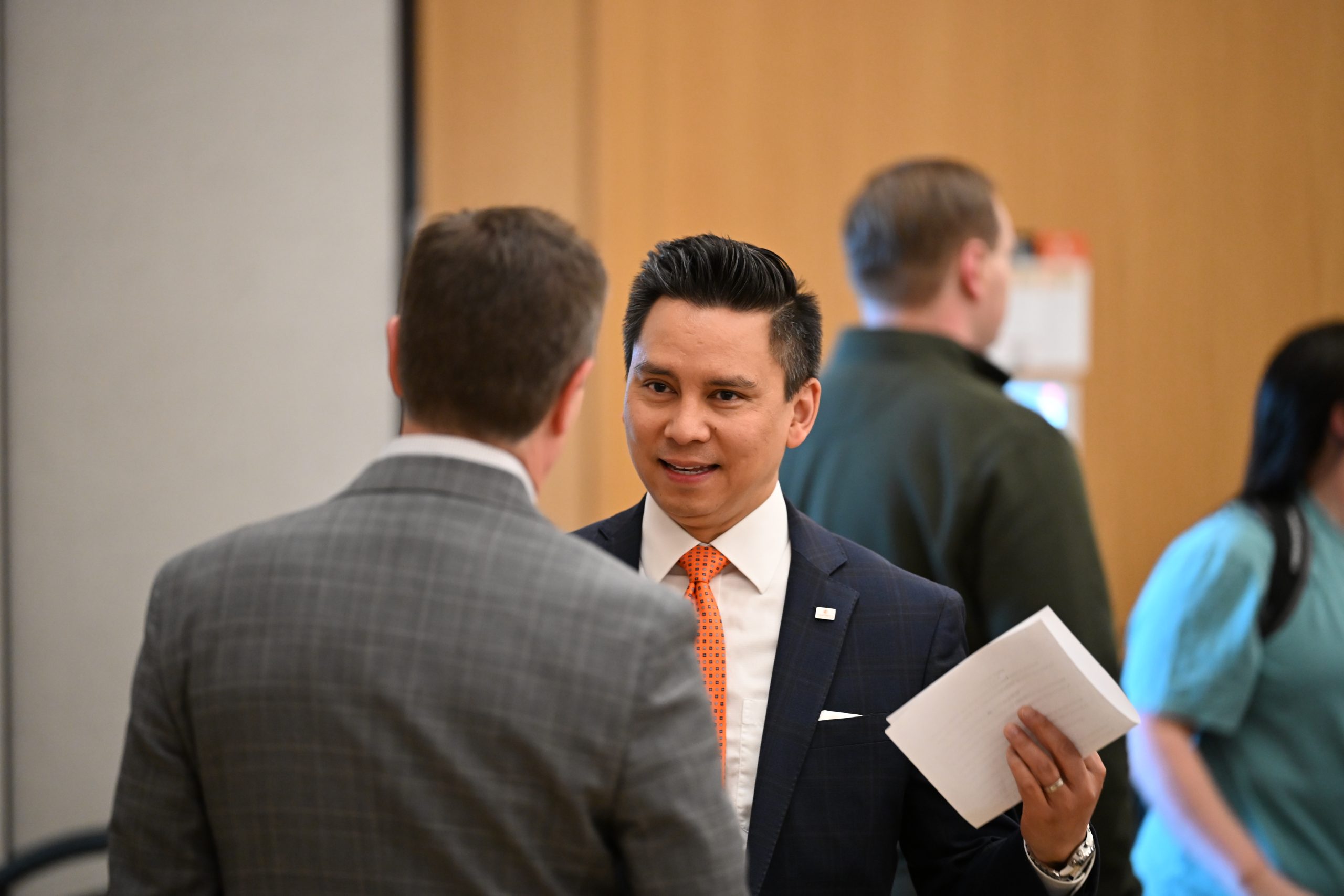 Man wearing a dark suit and orange tie holding a sheet of paper and talking with another man