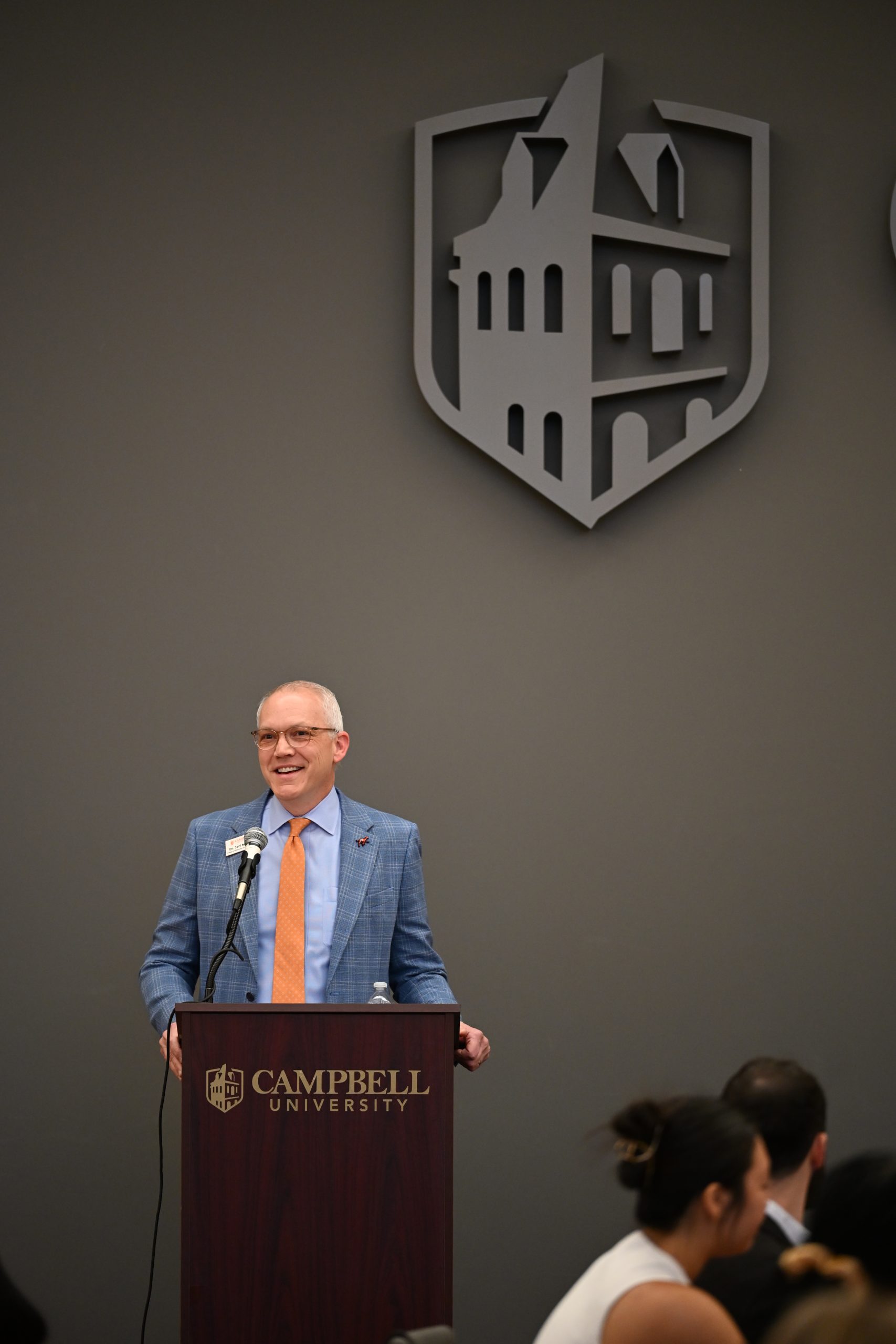 Man standing a podium wearing blue suit with blue shirt and orange tie