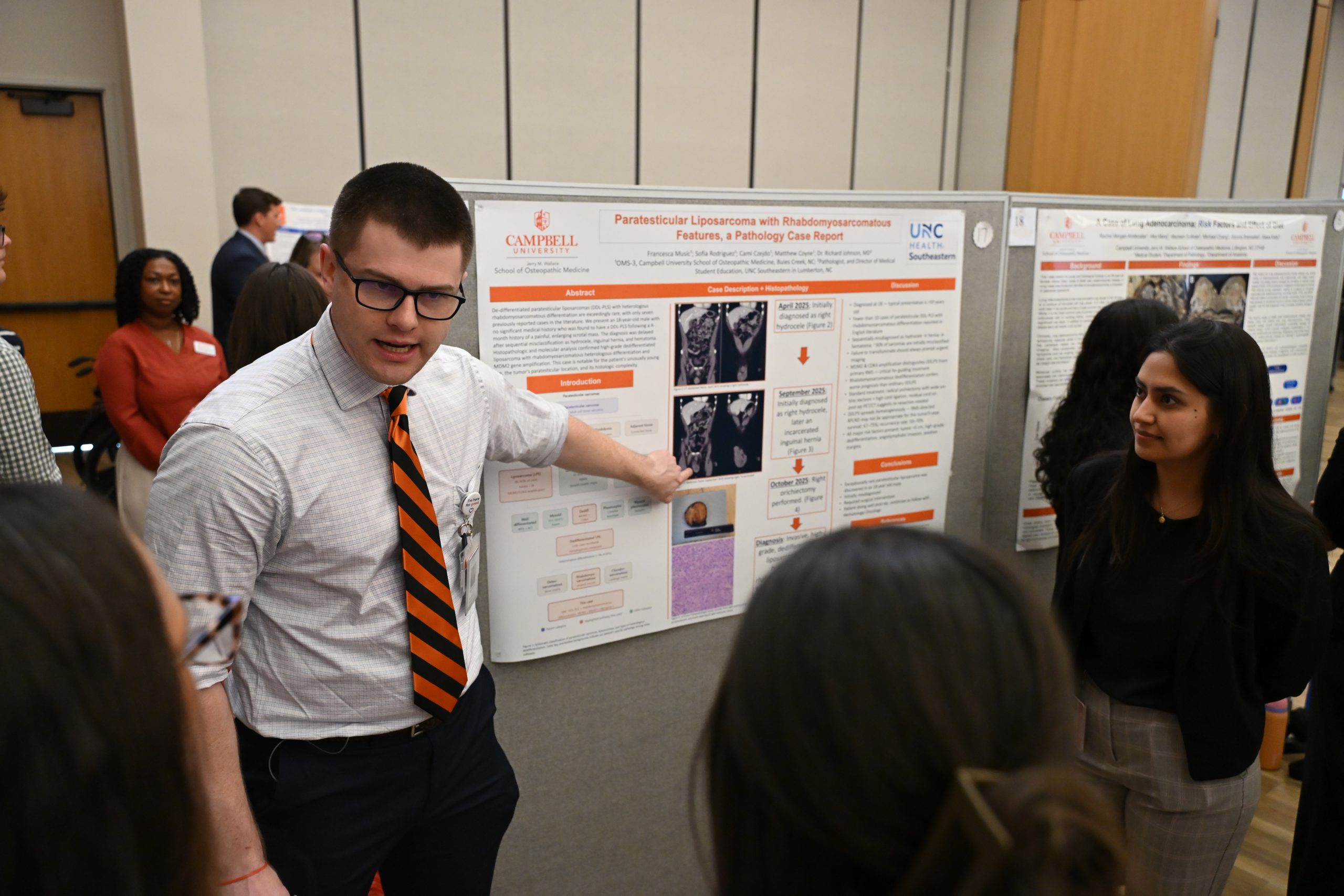Male student in shirt and tie pointing to poster and explaining research to attendees