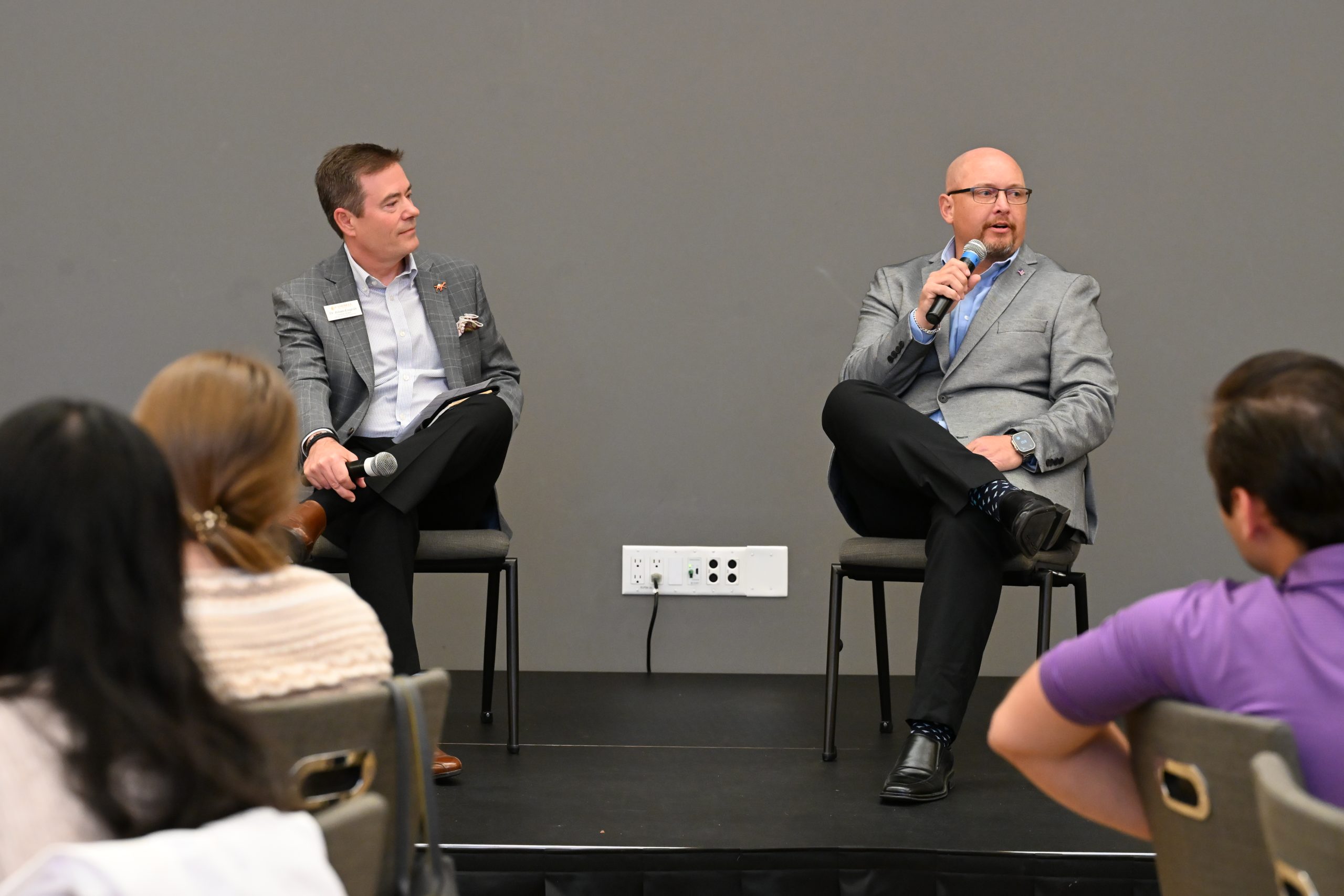 Two men sitting on stage wearing grey blazers and black pants holding microphones