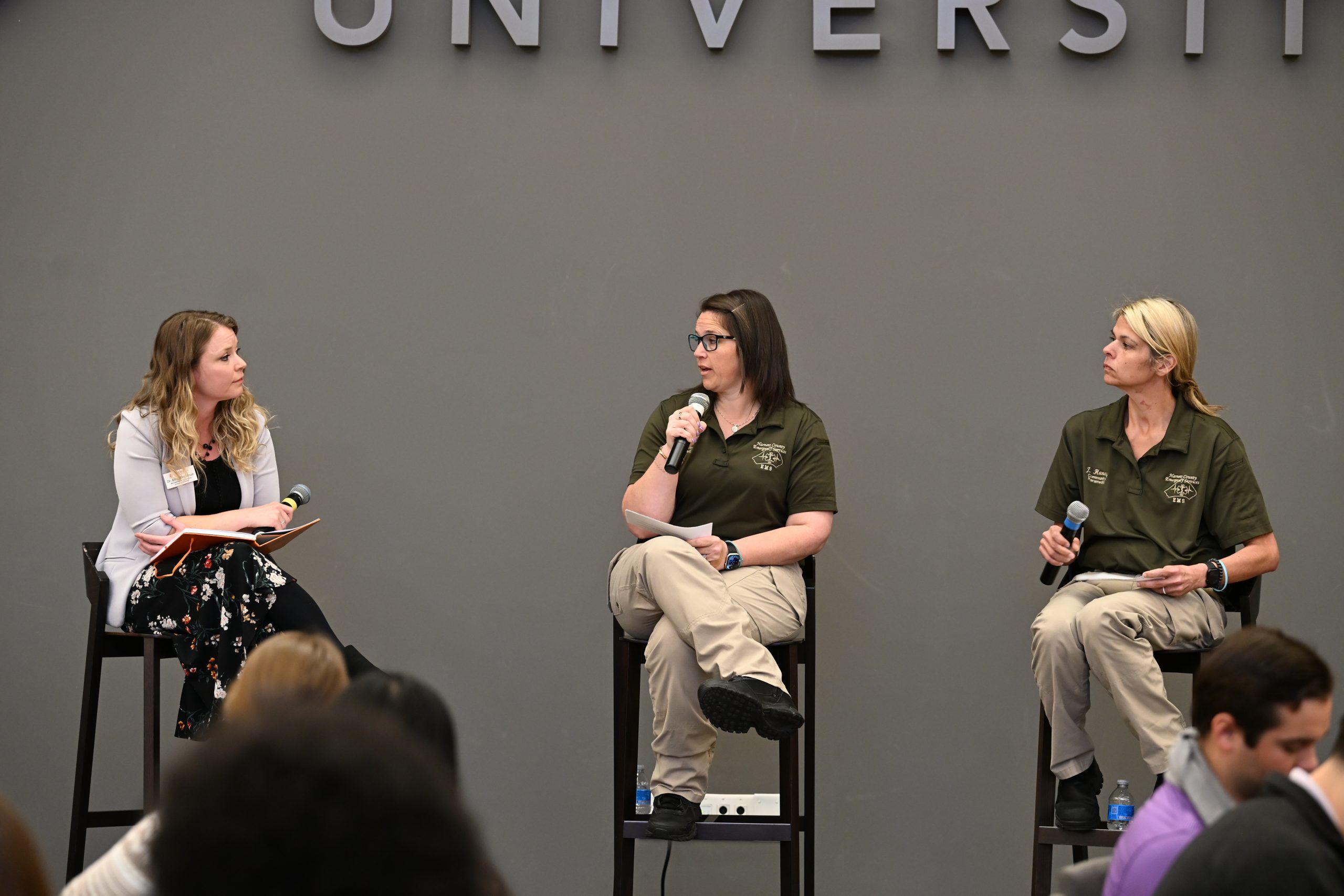 Female professor holding microphone sitting on a bar stool on stage with two community paramedics wearing green shirts and tan pants holding microphones