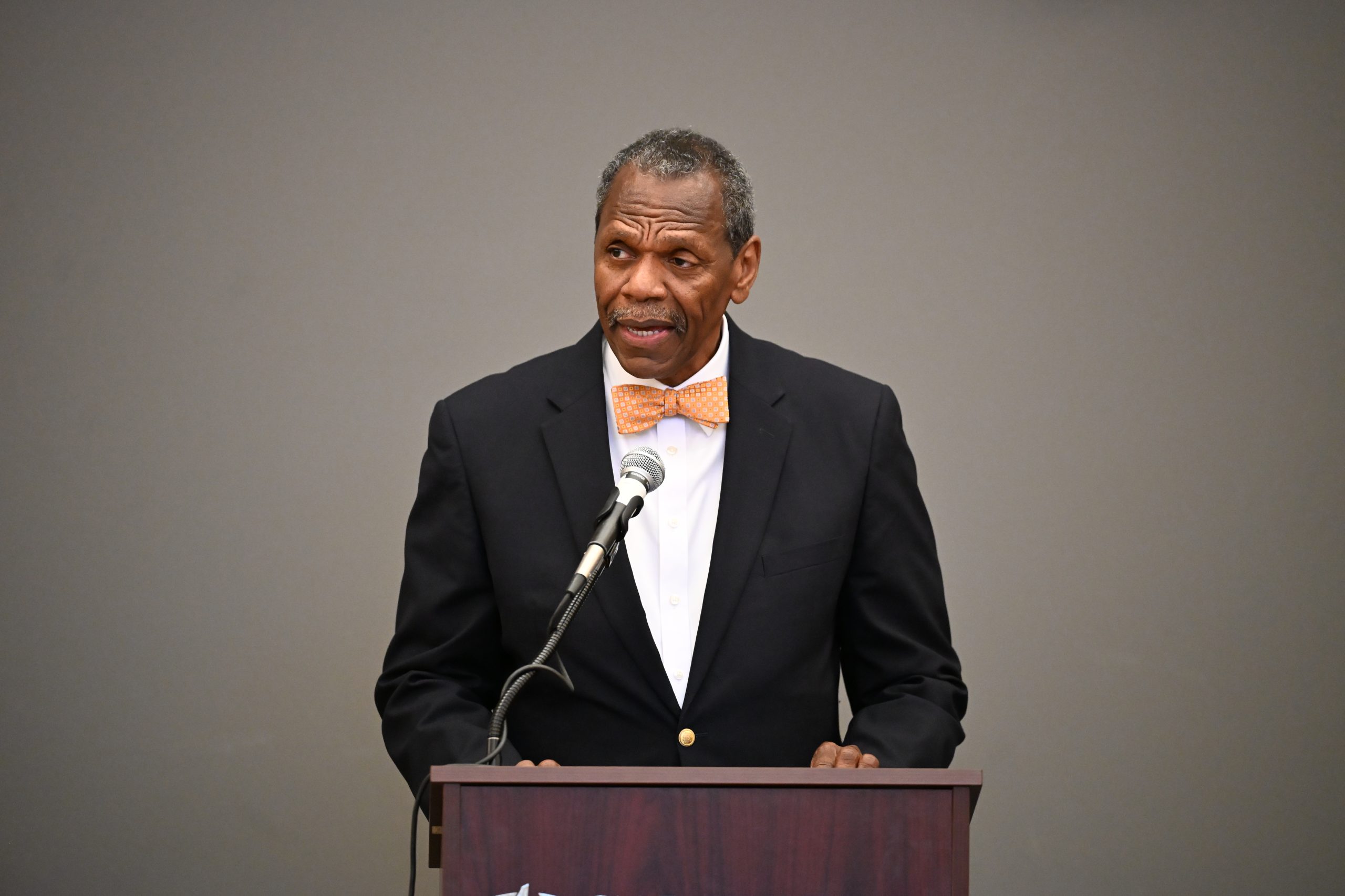 African-American male wearing black blazer and orange bow tie standing at podium