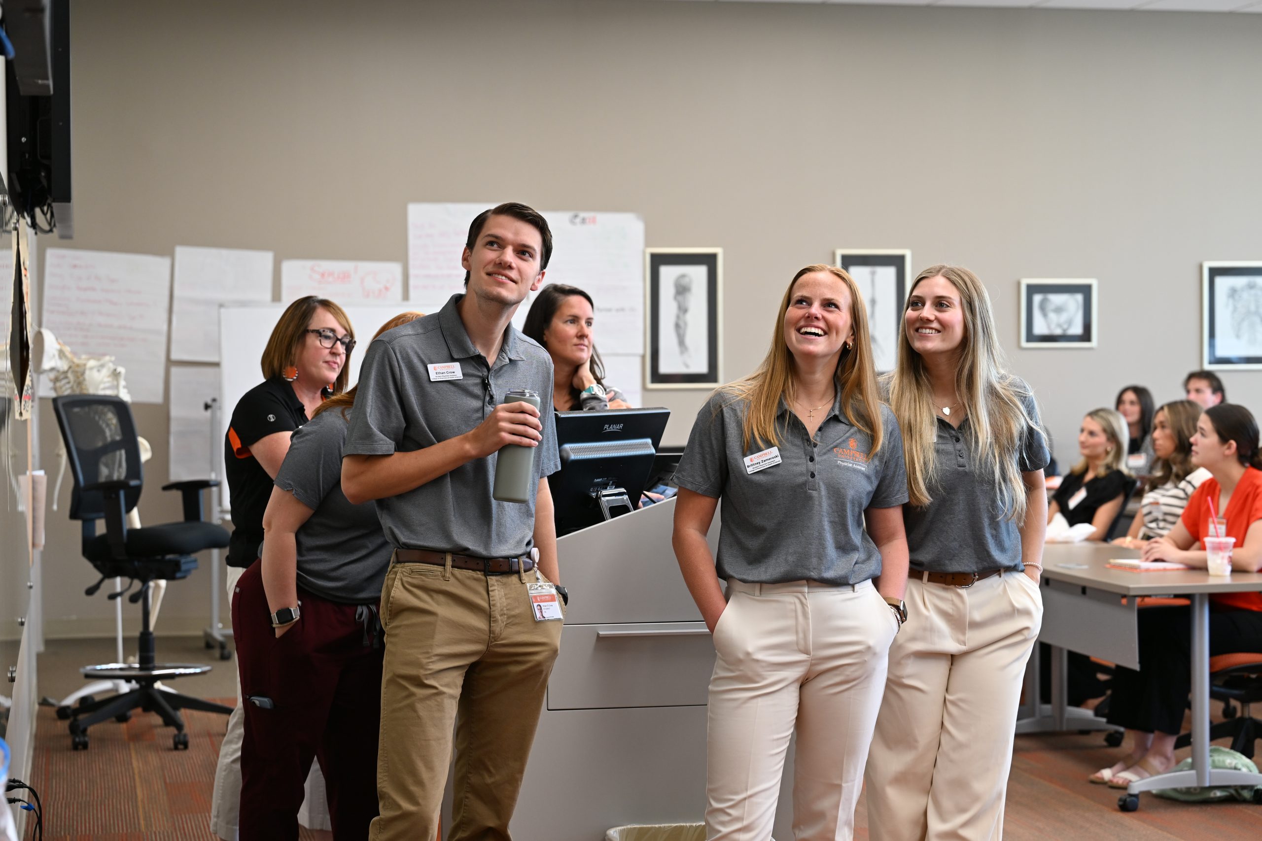 male and female PA students wearing grey polos looking up at screen in classroom with big smiles