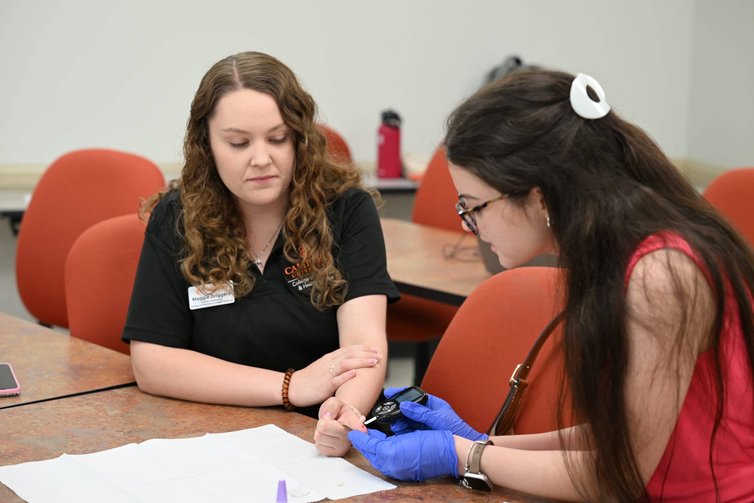 Female pharmacy student wearing black polo shirt holds out finger for prospective female student to test blood glucose in an activity.