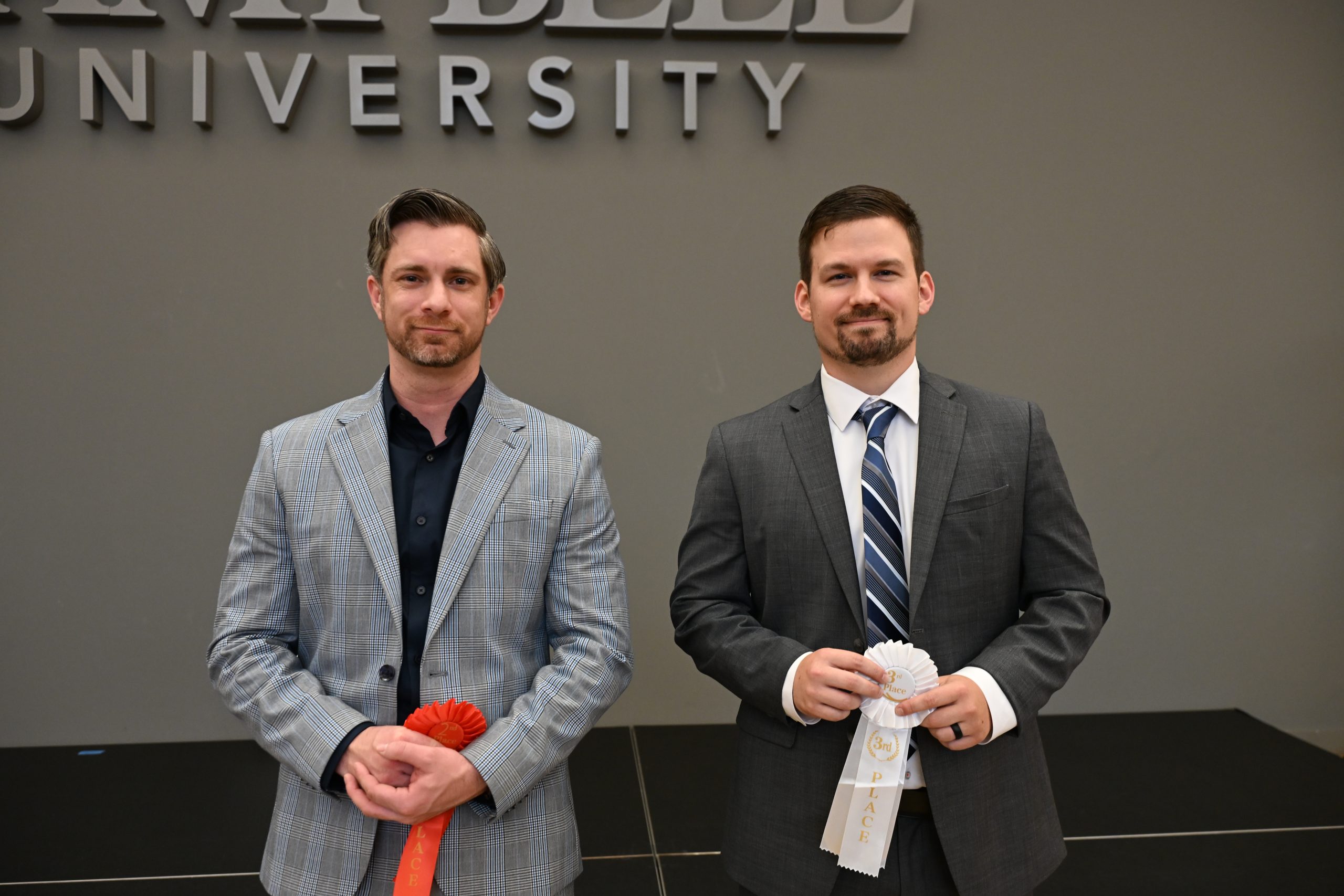 Two professionally dressed male students holding award ribbons