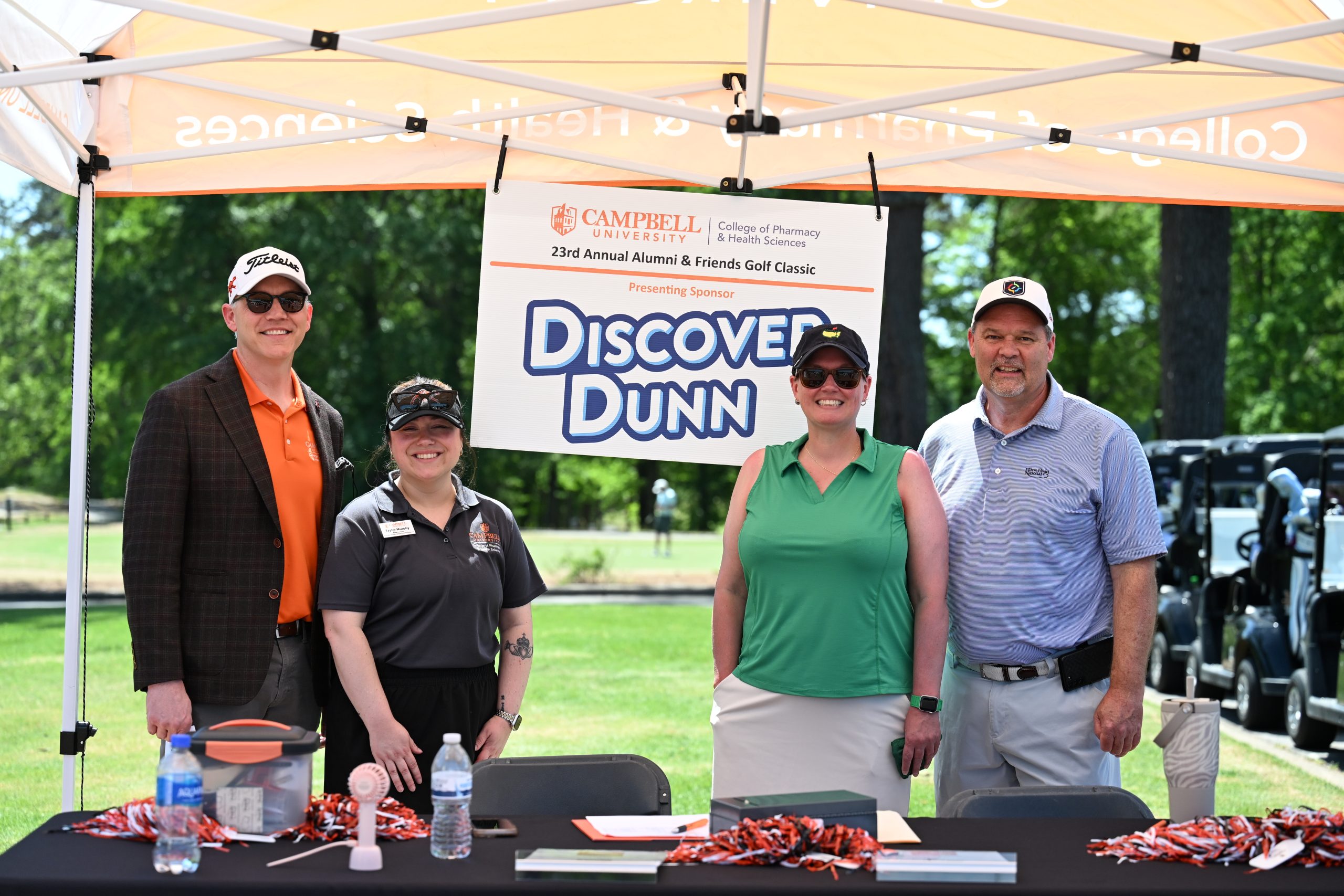 Two males and two females stand with presenting sponsor sign under tent