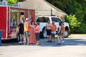 A group of people standing outside at a food truck. Several people are wearing orange and appear to be in conversation.
