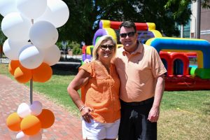 Two people pose for a photo next to a stand with white and orange balloons and an inflatable multi-colored obstacle course in the background.