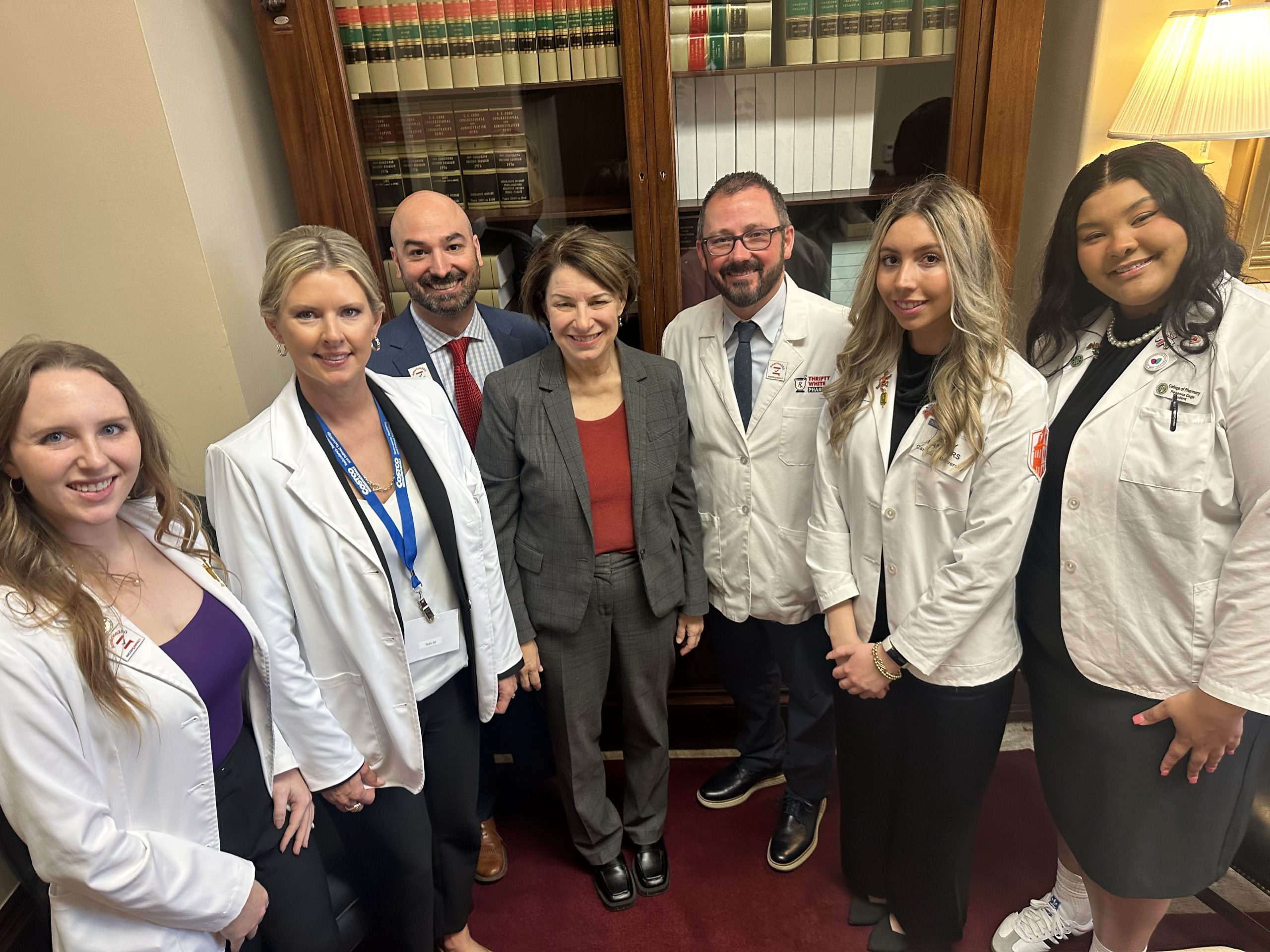 Pharmacy students wearing white coats standing with two members of Congress in an office in front of a bookcase