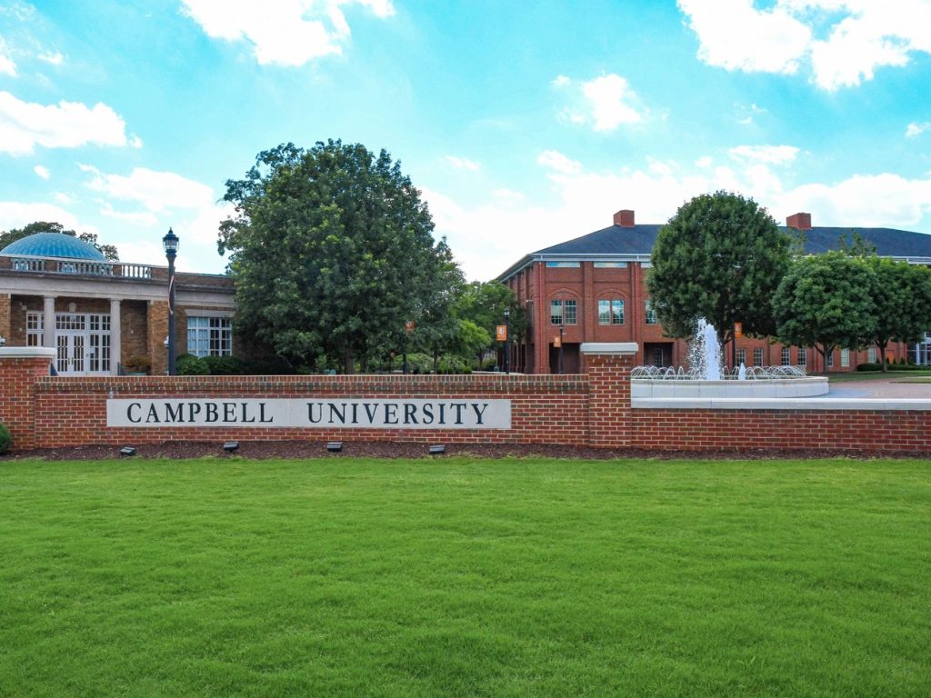 Campbell University sign in brick and concrete with a fountain behind it and with Carrie Rich Hall and Maddox Hall in the background