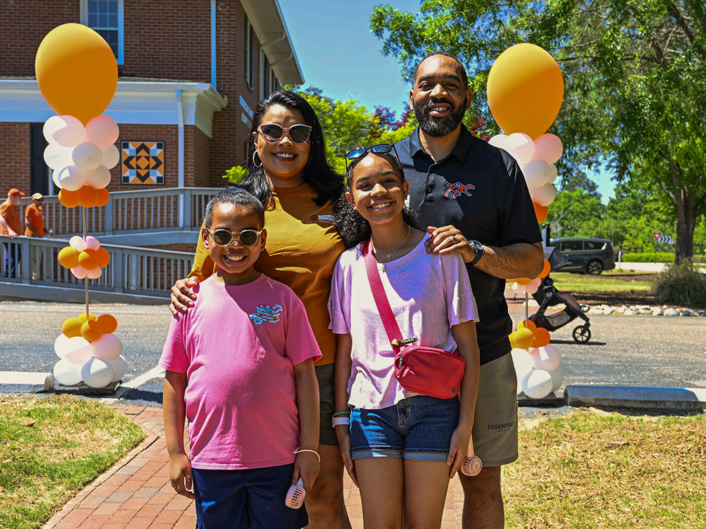 A family of four pose for a photo, smiling and outside. Orange and white balloons are in the background along with a brick building and a leafy tree.