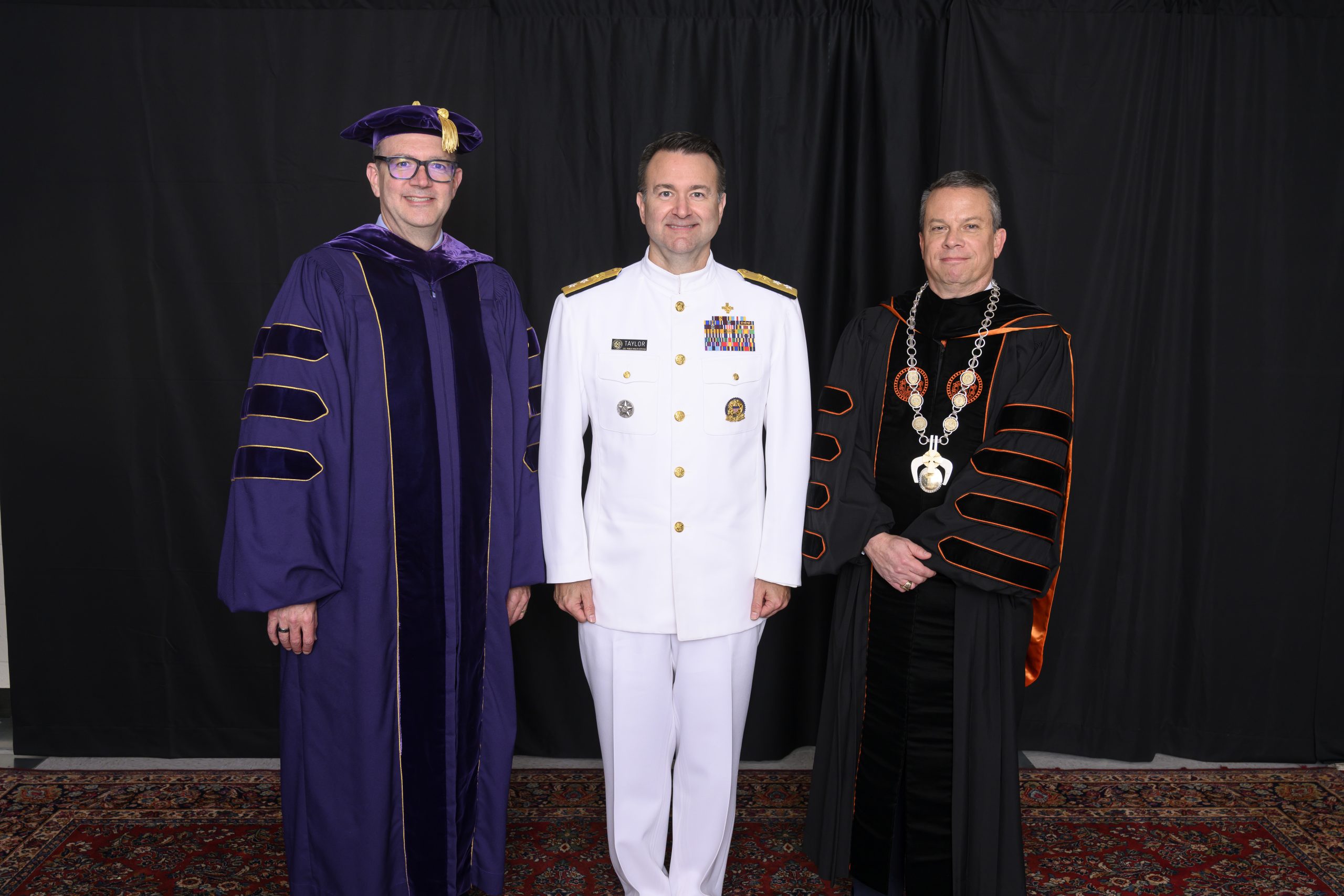 Male wearing purple regalia on left, male wearing white formal military uniform in the middle, and male wearing regalia