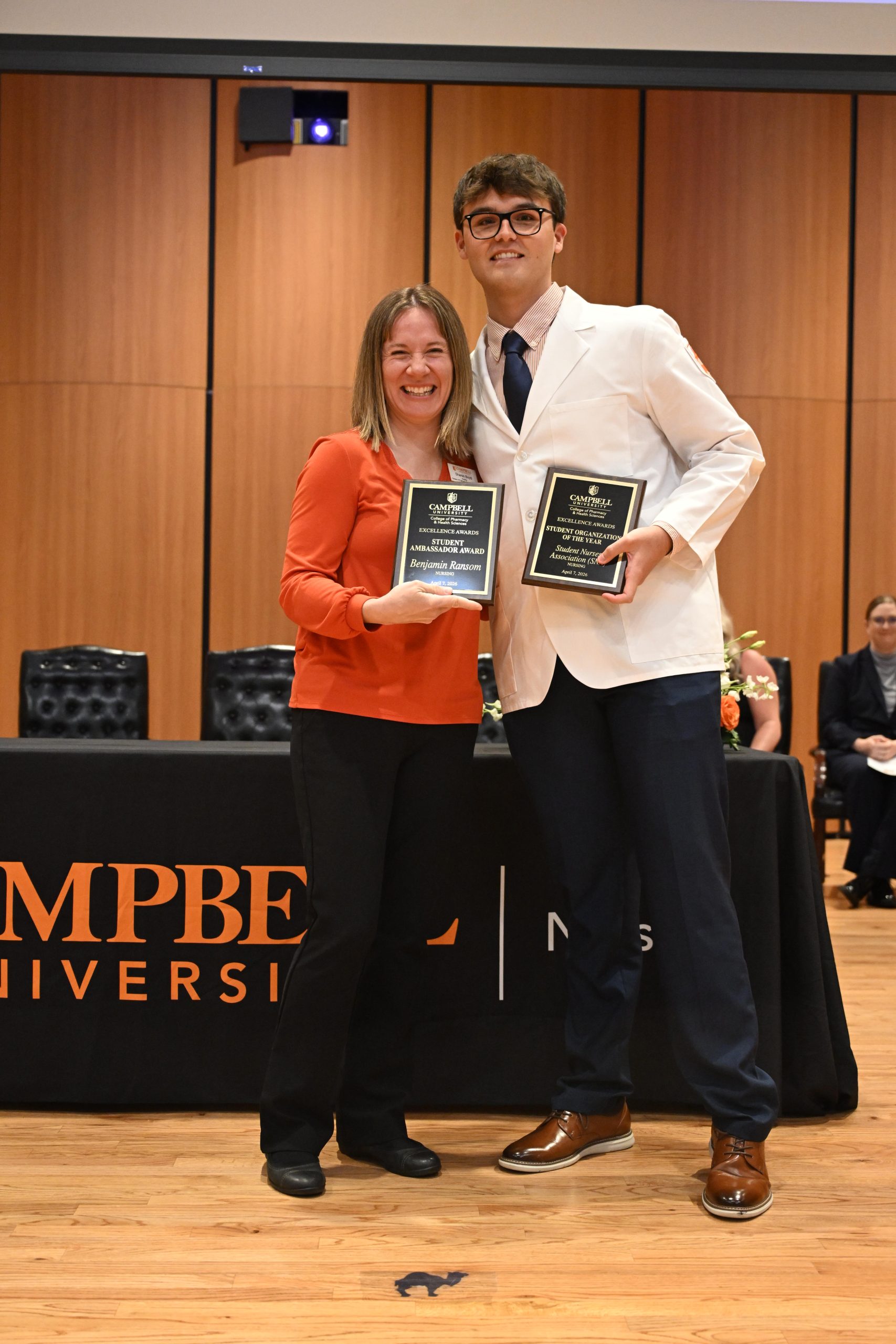 Female faculty standing with male student holding two plaques.