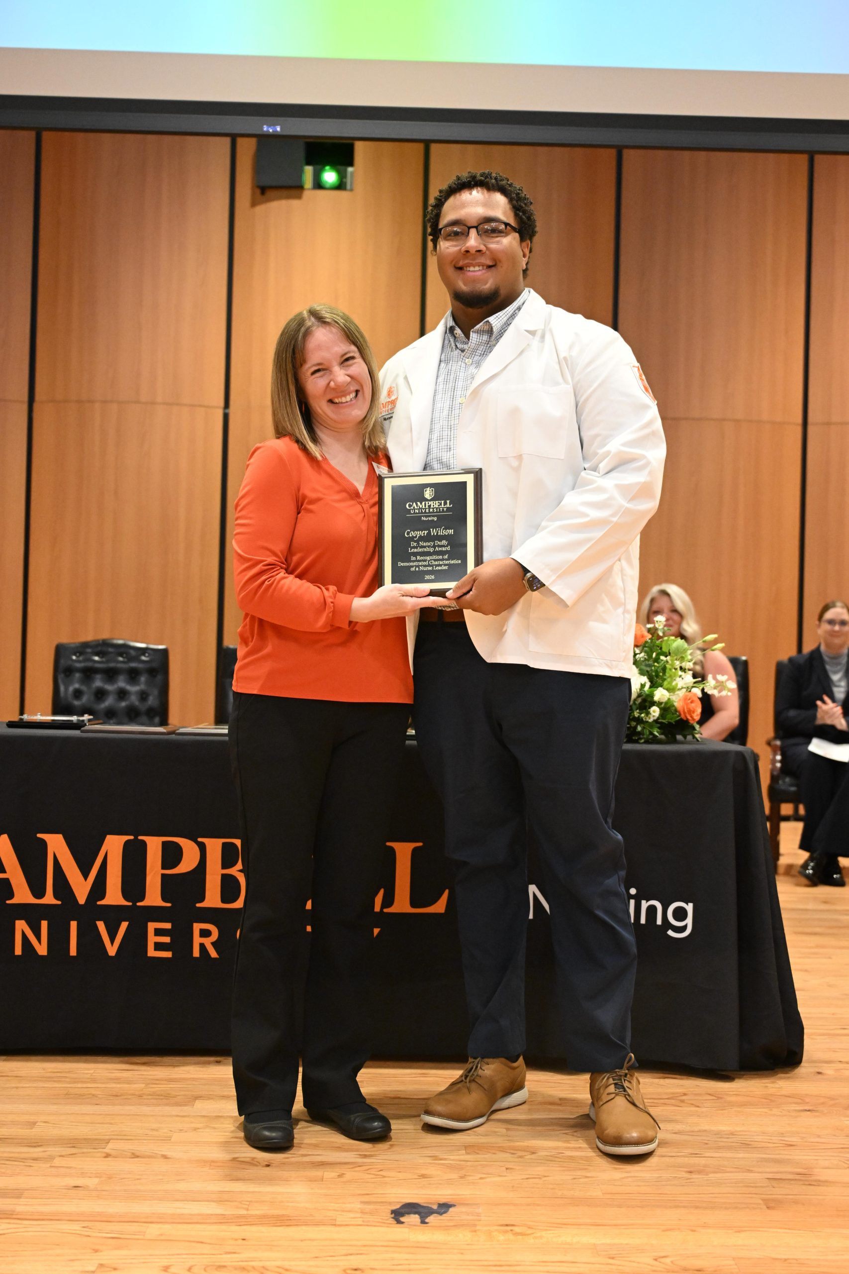 Female faculty member wearing orange shirt stands with male student holding a plaque.