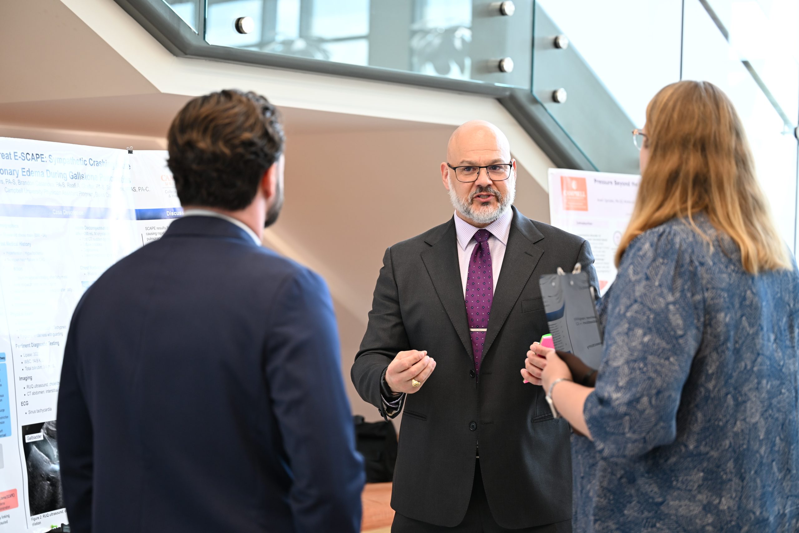 Man in suit talkking about his poster presentation to a female faculty member and another male student