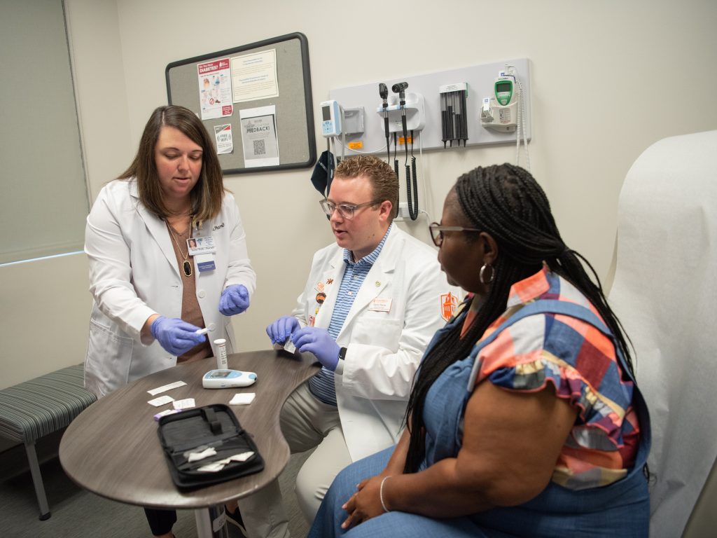 Female pharmacist in white coat assisting male student pharmacist in white coat who is sitting beside a female patient.