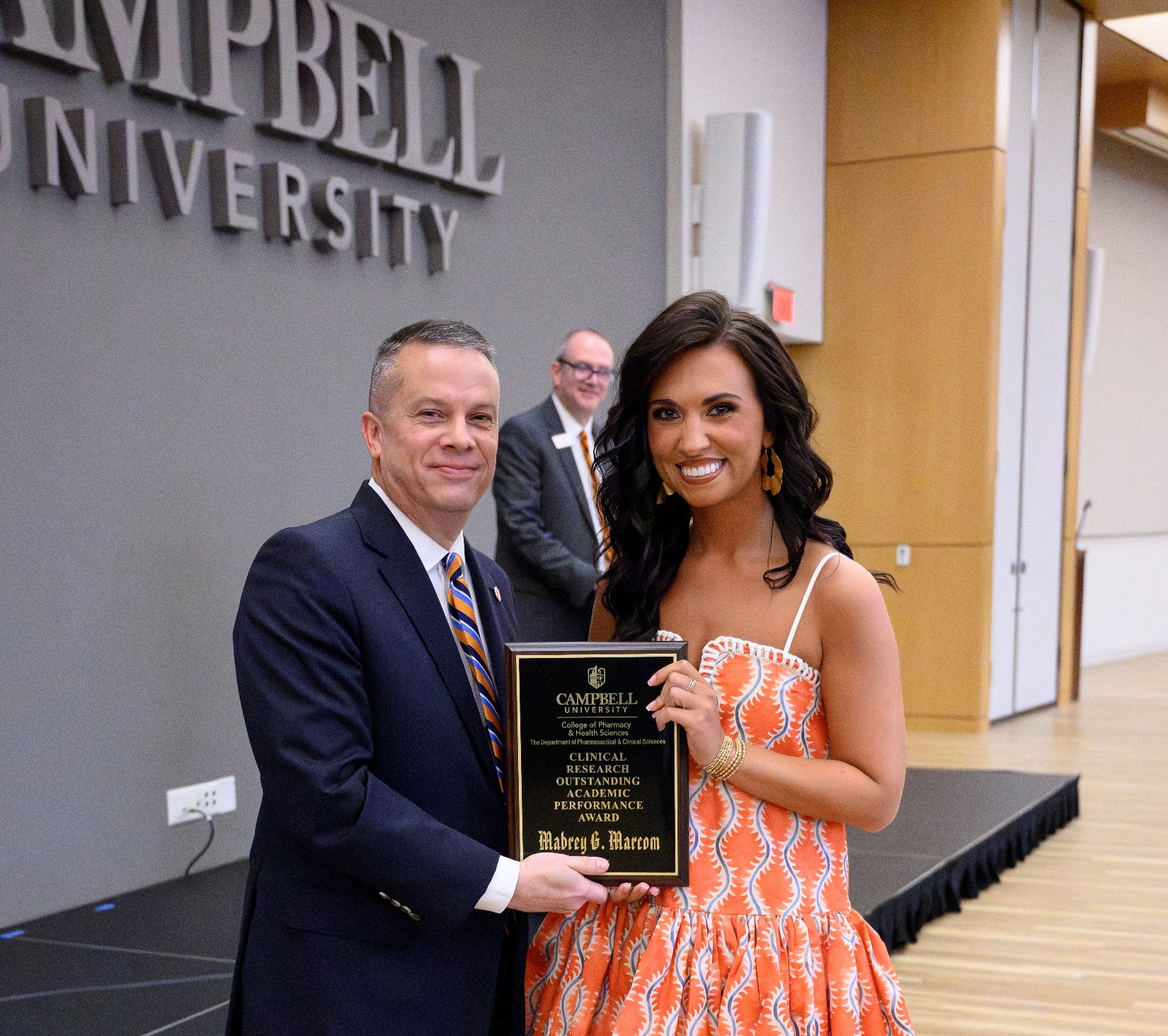 Male in dark suit standing beside female in orange dress both holding a plaque