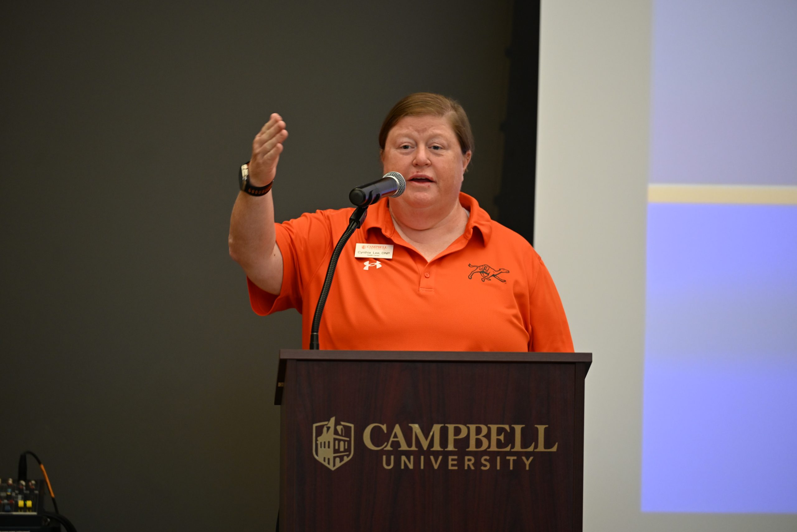 Dr. Cynthia Lee wearing an orange Campbell shirt holder her hand up gesturing while at the podium.