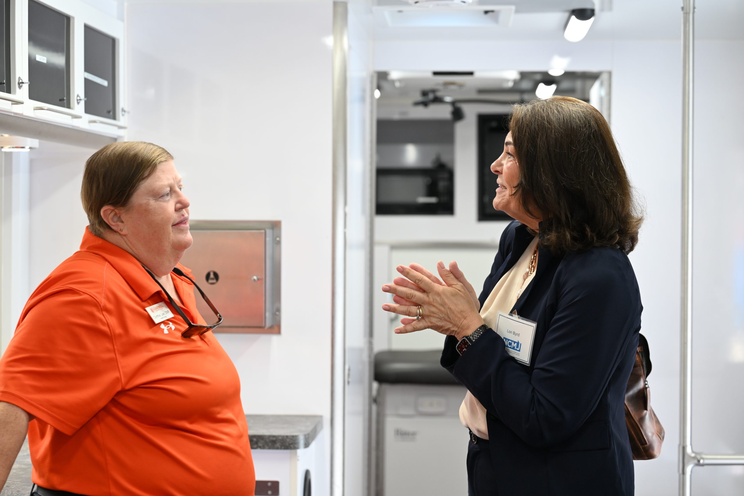 Two ladies talking. Women on left in orange shirt and women on right in navy blue suit.