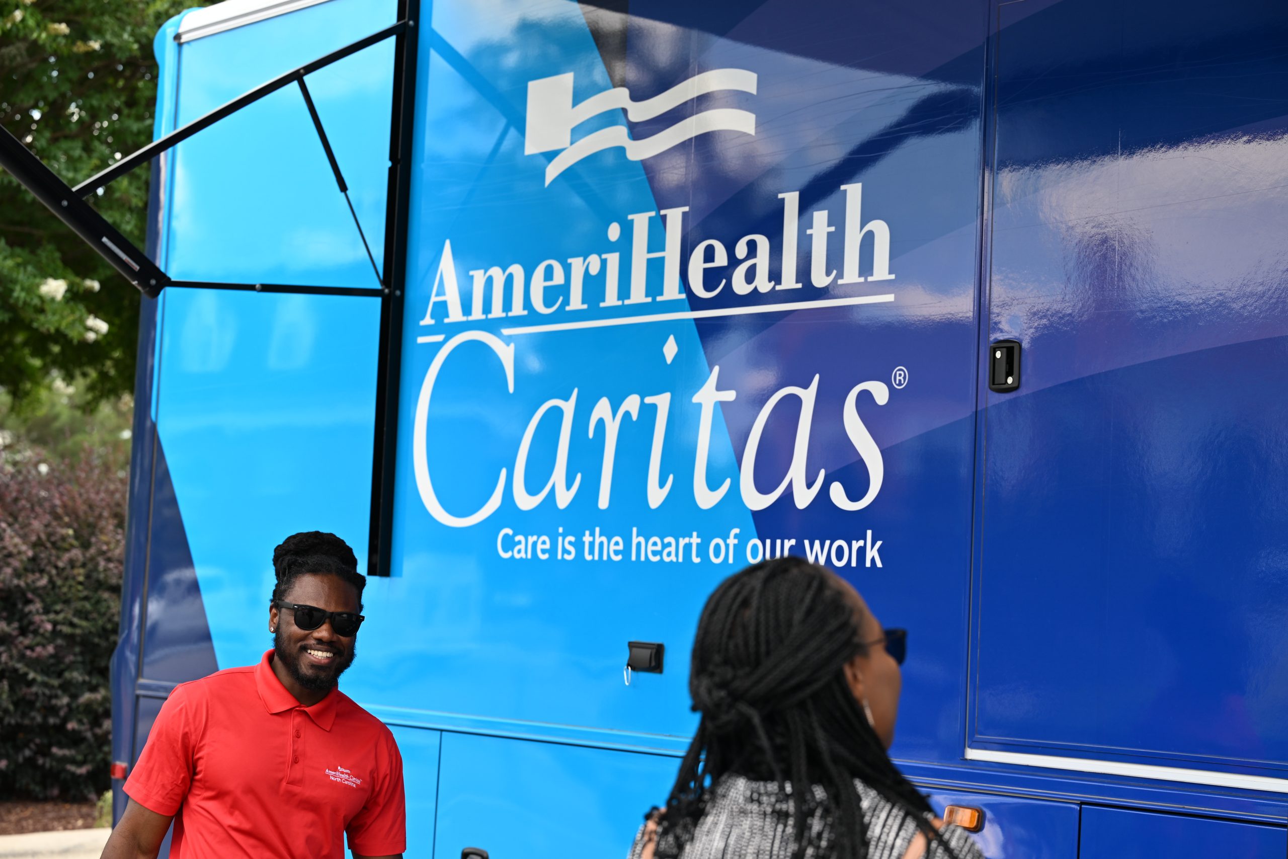 Blue AmeriHealth Caritas mobile healthcare vehicle with man wearing sunglasses and orange shirt standing beside it smiling.