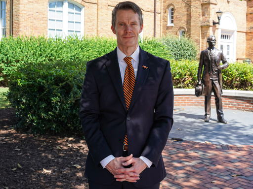 A person in a suit stands outdoors on a brick path, with a statue of J.A. Campbell is in the background. The scene includes green shrubs and a brick building.