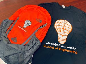 An orange and gray backpack sits on top of a black t-shirt. Both the backpack and t-shirt have "Bob Barker Engineering Camp of Future Innovators" written inside a lightbulb design. Below that is text that says "Campbell University School of Engineering"