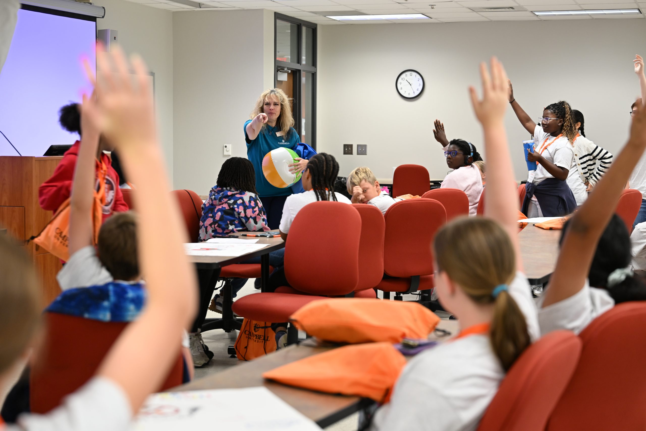 Female teacher holding beach ball points to students raising their hands
