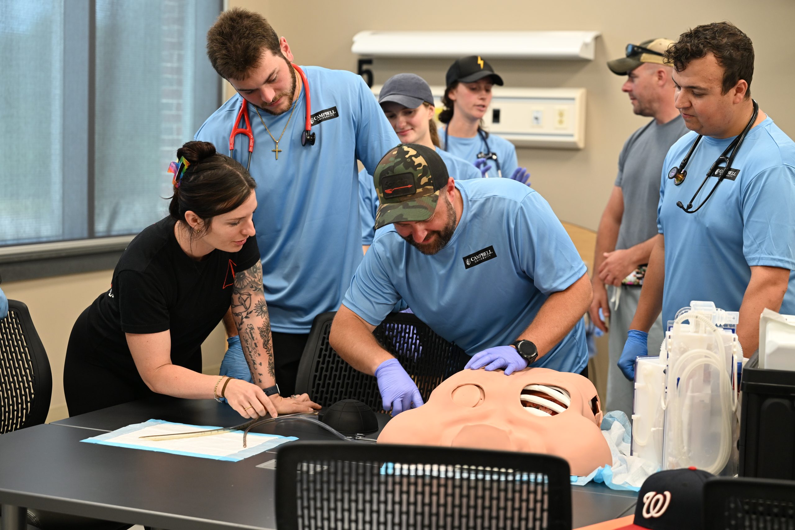 Male PA student learning how to insert a tube into a lung with two male PA students watching while a female teacher instructs