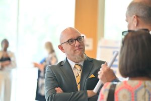 A man in a gray suit with a yellow tie listens attentively while speaking with attendees during a research poster session at the Campbell University College of Pharmacy & Health Sciences.