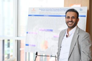 A smiling presenter stands beside a research poster display during a research symposium at the Campbell University College of Pharmacy & Health Sciences.