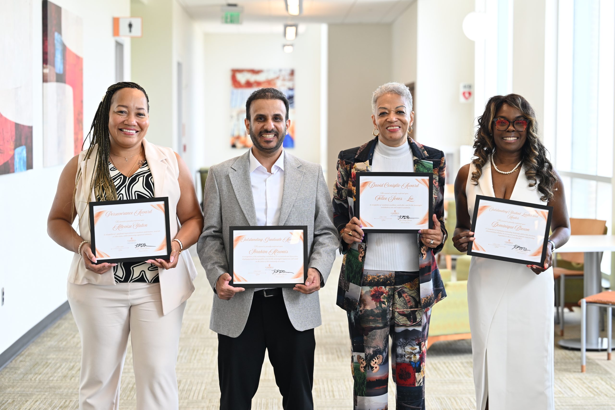 Four award recipients stand side by side in a bright hallway at Campbell University College of Pharmacy & Health Sciences, each smiling while holding a framed certificate. From left to right: a woman in a cream pantsuit, a man in a light gray blazer, a woman in a colorful patterned suit with a white turtleneck, and a woman in a white sleeveless dress. The group faces the camera in a posed recognition photo with large windows and campus artwork visible in the background.