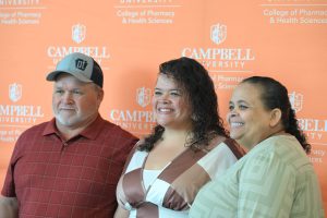 Three people smile while posing together in front of an orange Campbell University College of Pharmacy & Health Sciences step-and-repeat backdrop during a recognition event. A young woman stands between two adults, and all three face the camera.