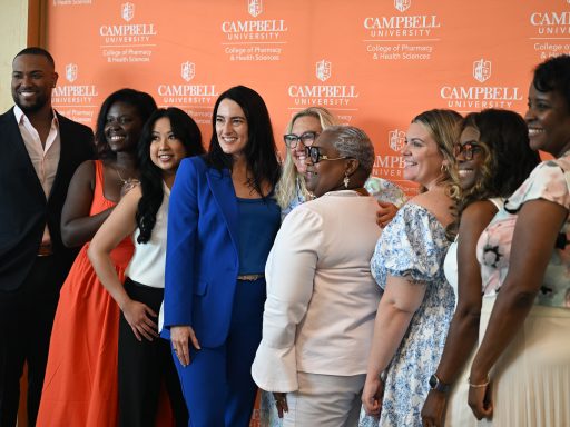 A diverse group of students gather in front of a Campbell University College of Pharmacy & Health Sciences backdrop for a group photo. A woman in a bright blue suit stands in the center, surrounded by smiling attendees in professional and business-casual attire.