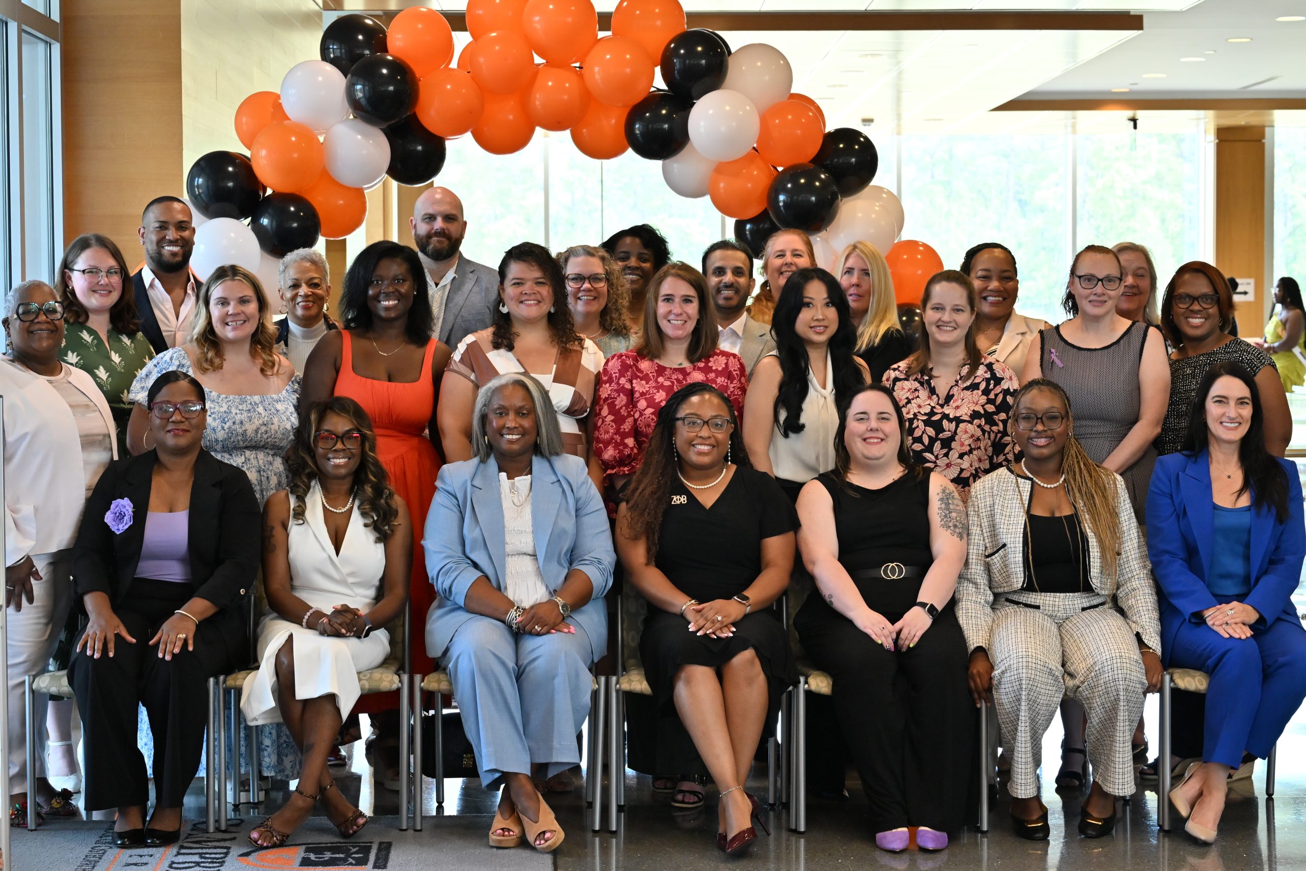 Students pose for a large group photo beneath an orange, black, and white balloon arch inside the Campbell University College of Pharmacy & Health Sciences. The group is arranged in seated and standing rows and smiles toward the camera.